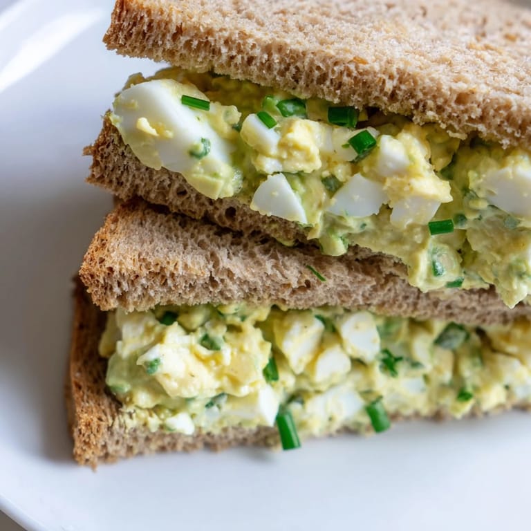 A close-up of avocado egg salad sandwich stacked with sliced tomatoes and chives on rustic bread.